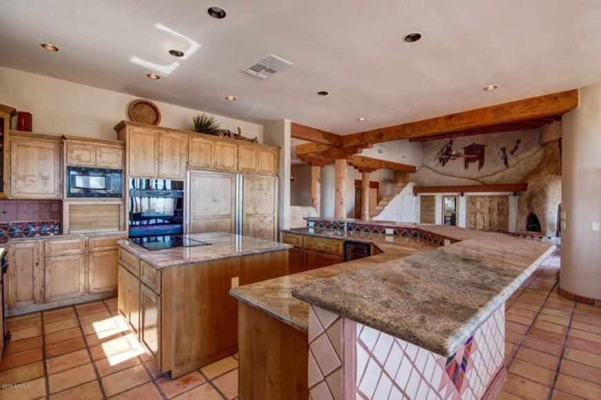 西南厨房，带一个早餐岛，周围是木制橱柜和岛式吧台，台面在石板地板上凸起。