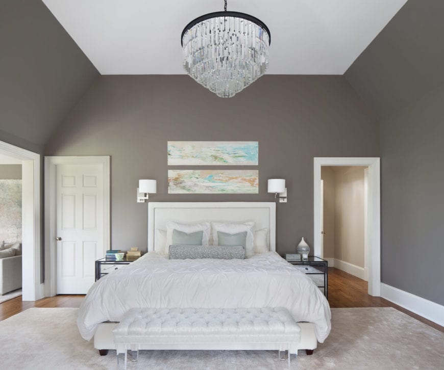 白色天花板上的水晶吊灯在灰色墙壁的映衬下格外引人注目。传统床的白色软垫床头板上挂着几幅彩色抽象画，床脚有一张配套的白色软垫长椅。