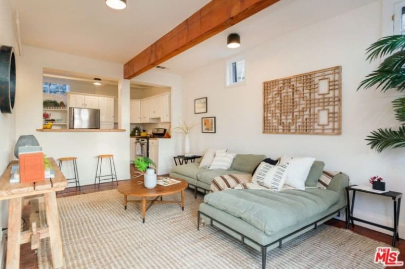 新鲜的客厅里有白色天花板上的木梁。它的设计是用木雕墙艺术安装在l型组合沙发上，沙发放在条纹地毯上。