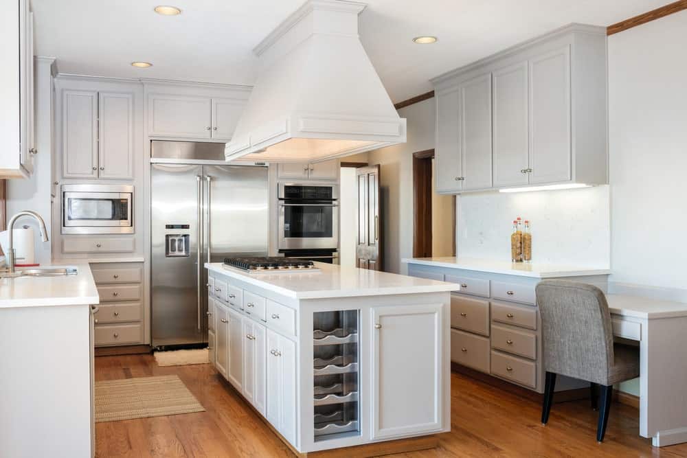 白色的厨房配以硬木地板，上面铺着棕褐色的地毯。它包括一个悬挂在早餐岛上的抽油烟机，内置灶台和用玻璃包裹的酒架。