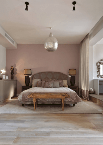 一个巨大的银色吊坠和木制床头柜上的台灯照亮了这间主卧室。房间里有一张粉红色图案的床，床的一端有一张木凳，上面铺着浅色的木板地板。