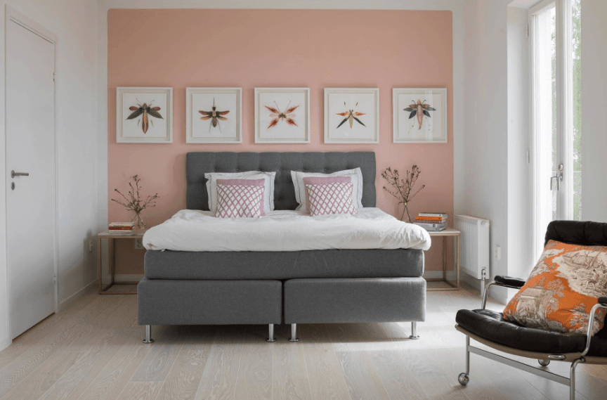 主卧室有淡粉色的强调墙，白色的墙壁艺术品安装在灰色的床上。它配有木制床头柜和带有橙色印花枕头的皮椅。