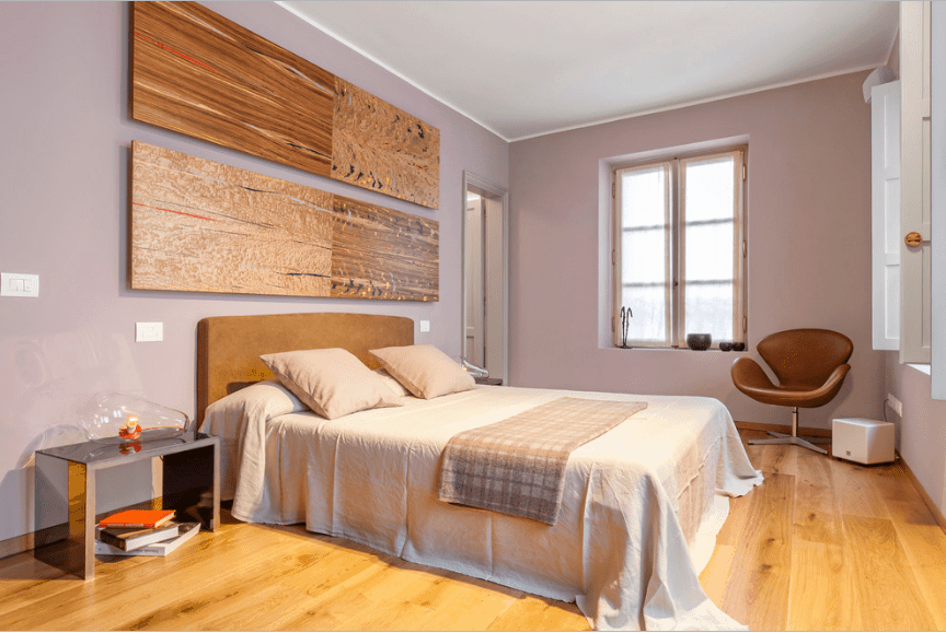 主卧室有裸粉色的墙壁和床上方的多面板木墙艺术。房间里有带镜子的床头柜，角落里有一把真皮椅子。