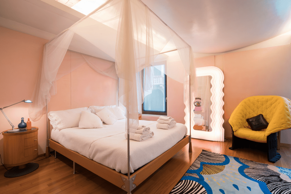兼收并蓄的主卧室配有木制顶棚床和床头柜，以及黄色的簇绒椅子和波浪形照明镜子。它有硬木地板，上面有蓝色印花地毯。