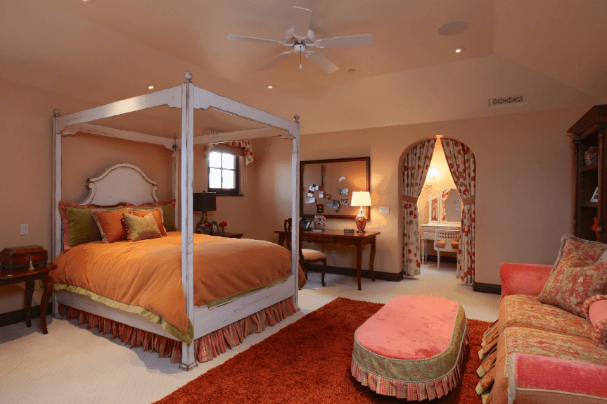 农家乐卧室，木质天篷床对着珊瑚粉色沙发，配有软凳。它包括一个覆盖着点状窗帘的开放式拱门，通往浴室的梳妆区。