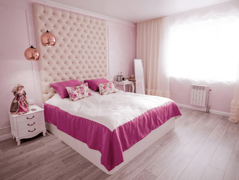 迷人的卧室拥有一个超大的浅粉色簇绒床头板安装在白色的床上。它被一对挂在白色床头柜上的铜吊坠照亮，上面有一个华丽的洋娃娃。