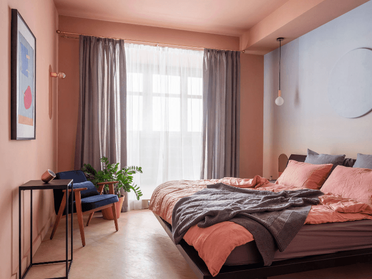 当代的主卧室，鲑鱼粉色的墙壁和灰色的窗帘与床互补。它装饰着一面圆形无框镜子，黑色金属桌子上方挂着一件墙壁艺术品。