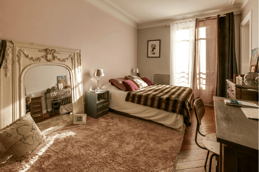 在这间主卧里，浅粉色的墙上挂着一面巨大的、华丽的白色镜子。它的旁边是一个灰色的床头柜，上面有一个蓬松的地毯覆盖着木板地板。