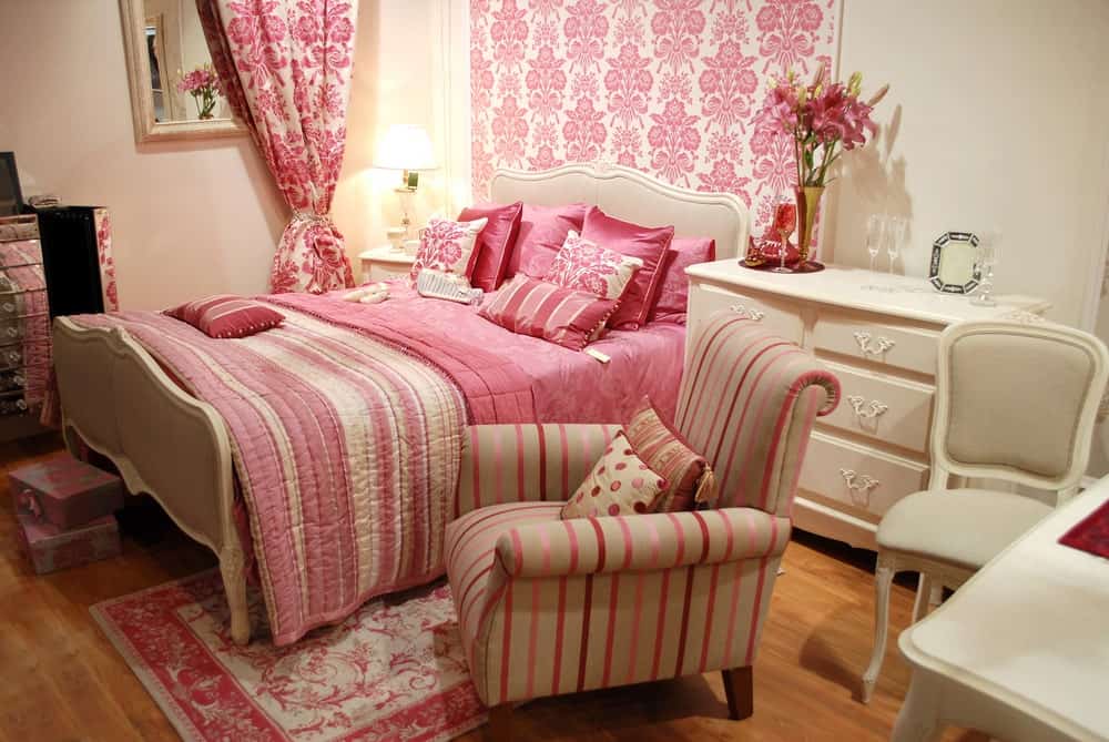 迷人的主卧室与粉红色的碎花壁纸相配的窗帘。房间里有一张软垫床，还有一把条纹椅子，椅子放在硬木地板上的粉红色地毯上。