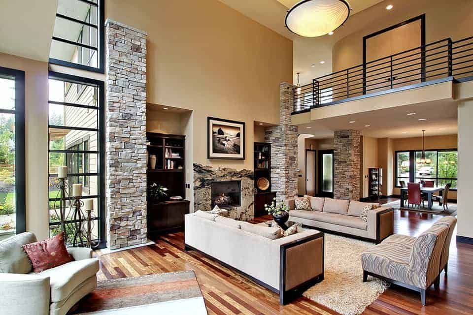 这间宽敞的客厅的米色墙壁以大石柱为特色，与壁炉的石面相匹配。一个带有黑色金属栏杆的室内阳台俯瞰着整个空间。