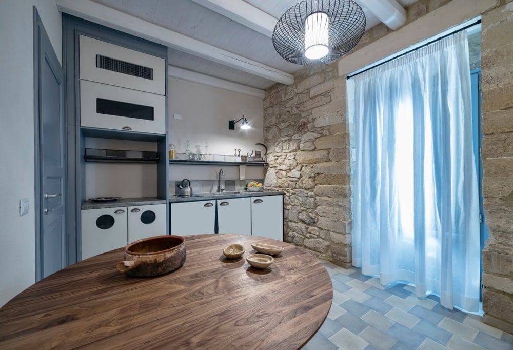 这是一个简单的西南风格的厨房，有一个漂亮的粗糙纹理的石墙，支撑着这个厨房的大窗帘入口通道。这种有质感的墙壁与现代白色和灰色的小厨房半岛形成鲜明对比，后者与白色木制天花板和砖地板搭配得很好。