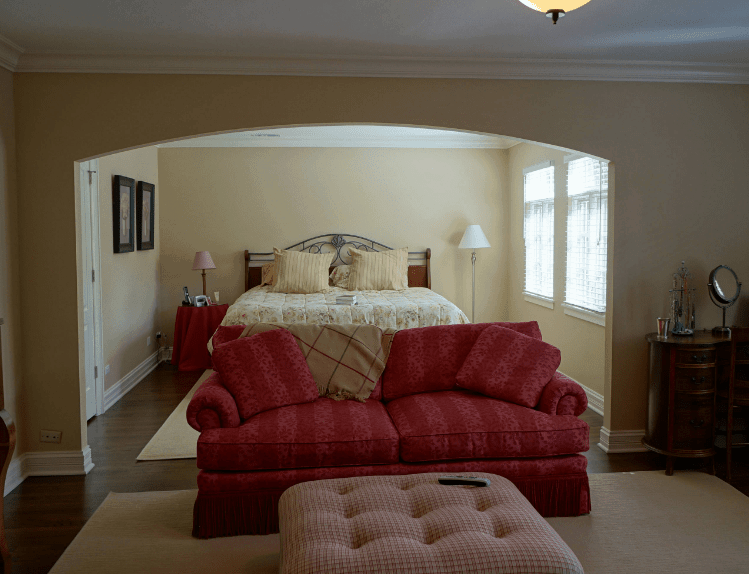 这间西班牙卧室的米色墙壁和地毯赋予它中性的色彩，与床脚的沙发和覆盖边桌的亚麻布的鲜明颜色相矛盾。平衡它们的元素是地板、橱柜和壁挂式框架的深色木色以及窗户的自然光。
