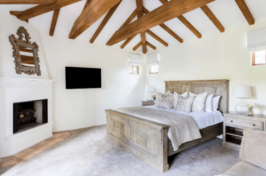 一个简单明亮的卧室，用浅色的色调来补充自然光。床架和床头柜的木质饰面与白色壁炉下的灰色地板和柔和的砖块完美搭配。这间西班牙式卧室的主要部分是拱形天花板，几根暴露的木梁形成了一个图案。