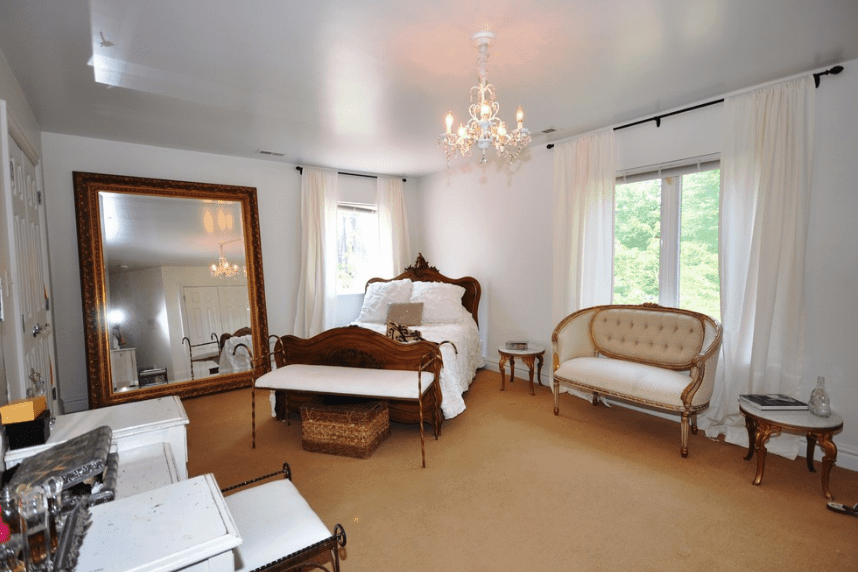 这是一间富丽堂皇的卧室，因为它宽敞的地板和黄铜镶边的大镜子高耸在床的一侧。墙壁和窗帘的白色给房间一种比实际更大的错觉。床、边桌和面粉的棕色元素补充了这种错觉。
