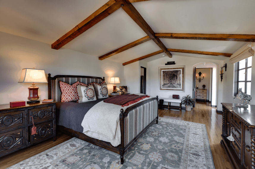 一间宽敞、天花板低、木梁外露的卧室。它的特色是一张可爱的风景照片，挂在白色床的一侧，黑色木床架和灰色条纹床头板。这与浅灰色的地毯和白色的墙壁很相配。