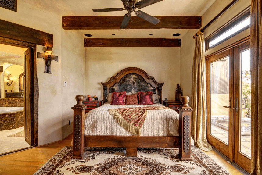 这是一间小而舒适的半凹室卧室，主要是一张带红木床边抽屉的木床。木材元素分散在玻璃门框、门拱和暴露的木制天花板梁中。来自玻璃门的自然光照亮了房间中央的图案地毯。