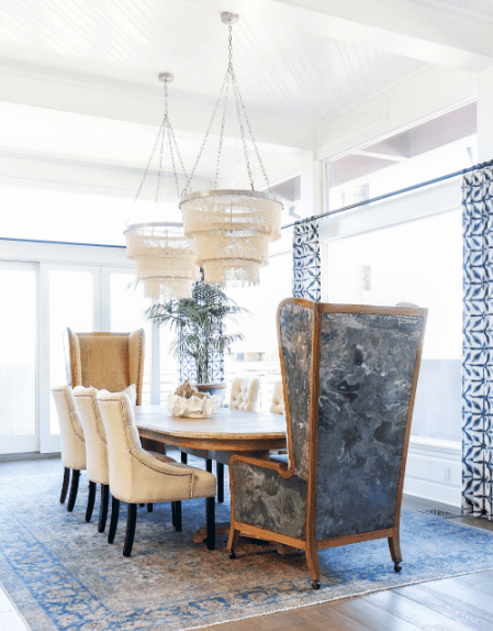 这是一个明亮的餐厅，依靠从窗户和玻璃射进来的自然光。白色的餐椅和从白色天花板上垂下的灯都装饰着它。这种明亮被窗帘、带图案的地毯和木桌头的高靠背椅的蓝色色调抵消了。