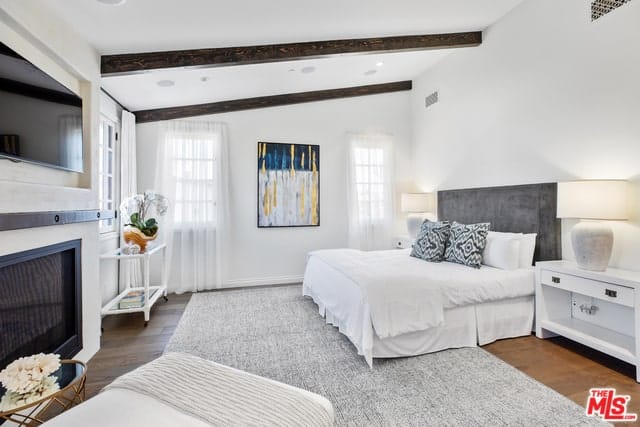 西班牙风格的卧室以暴露的木梁和木地板为特色，灰色地毯与白色的亮度形成鲜明的对比。它还突出了两扇落地窗中间的彩色抽象画。房间中央有一张白色的床，床头板是灰色的，正对着壁挂式电视，上面是一座现代化的壁炉。
