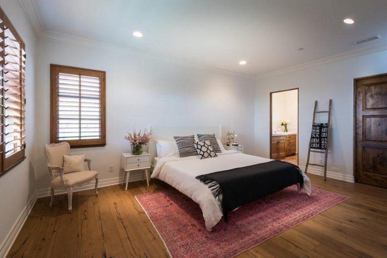 房间中间的粉红色地毯在这个中性白色的房间里是一个很好的亮点。然后通过各种各样的木制细节，如木地板、窗边、深色木门和用作毛巾架的乡村木梯来勾勒出这一点。一个优雅的法式椅子放置在角落作为一个不错的阅读角。