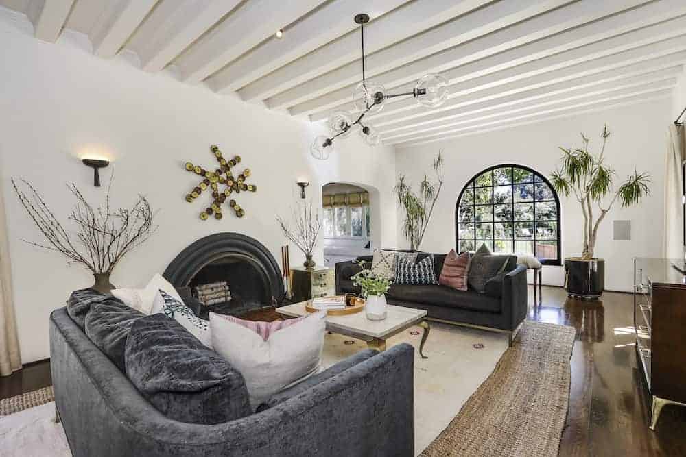 客厅有白色横梁天花板，白色灰泥墙，壁灯，壁炉，灰色沙发，硬木地板上的室内植物。