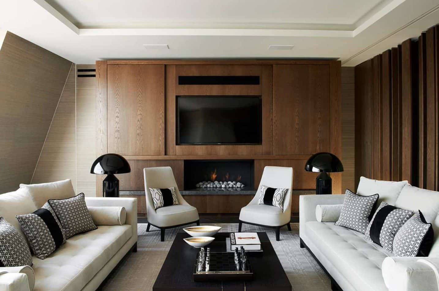 客厅里的现代壁炉和电视机前摆放着一对灰色椅子，黑色台灯照亮着椅子，客厅里摆放着簇绒沙发和深色木质咖啡桌。