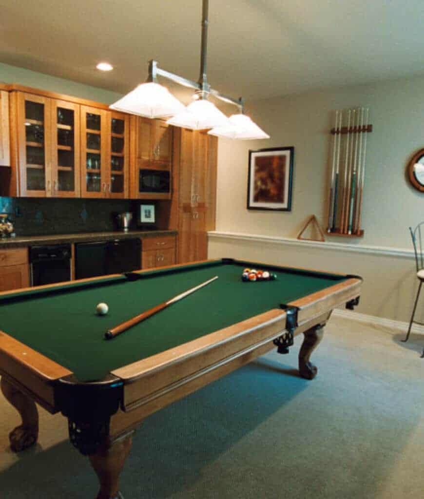 一个传统的台球桌，在吊灯下面的一侧有一个球杆墙架。房间内铺有地毯地板和内置橱柜，配有大理石台面。
