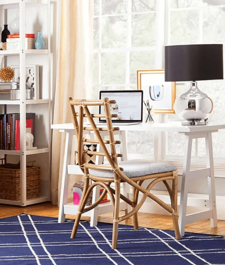 宽敞的家庭办公室里有一张白色的桌子和一张放在深蓝色地毯上的木椅。它由一盏黑色的台灯照亮，自然光从覆盖着桃红色窗帘的框窗中流过。