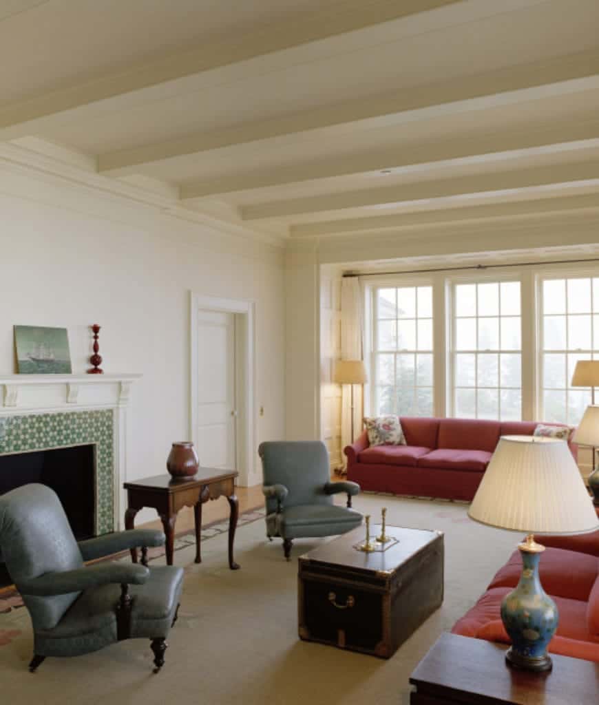在这间客厅里，一幅船墙艺术作品摆放在白色壁炉上，壁炉周围有绿色图案的瓷砖，客厅里有红色的隔断椅和灰色的扶手椅。