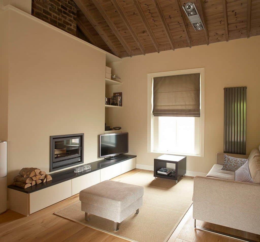 米黄色的客厅设有一座现代化的壁炉和一台放在木架上的电视。它有木梁天花板和宽阔的木板地板，上面铺着编织地毯。