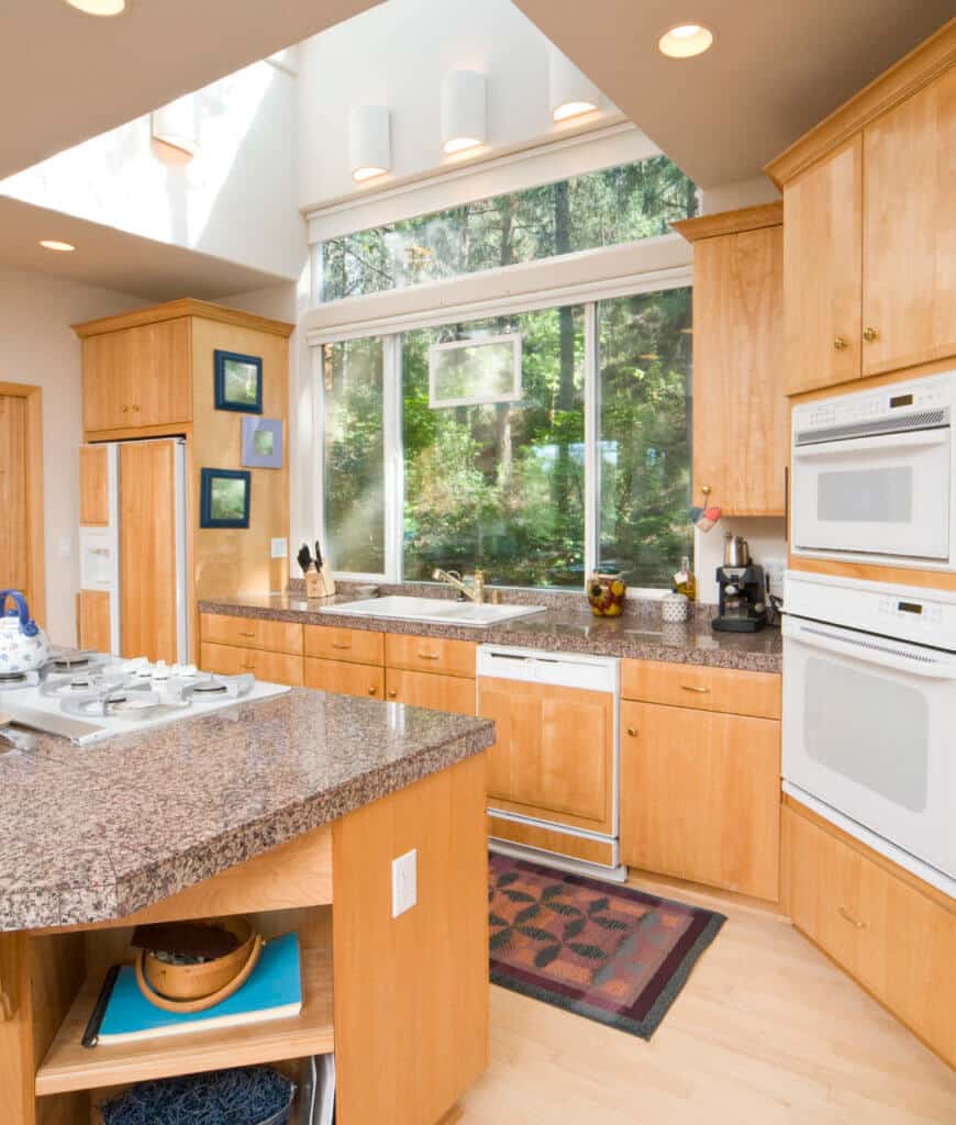 明亮的厨房，浅色硬木地板和玻璃镶板窗户俯瞰宁静的室外景色。它包括白色电器、木制橱柜和一个带内置搁架的大理石顶部厨房岛台。