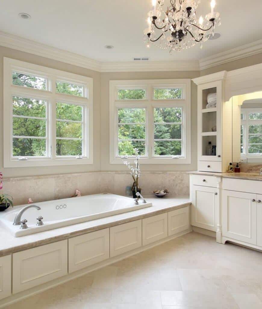这间主浴室拥有一盏华丽的吊灯和一个毗邻白色洗手台的浴缸，洗手台配有大理石台面和内置架子。