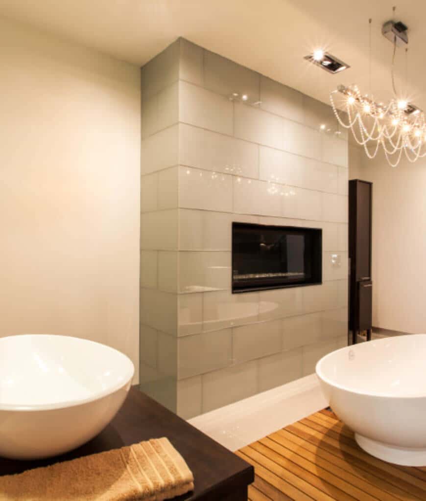 线性水晶吊灯照亮了这间主浴室，设有一个独立的浴缸，浴缸位于木甲板上，面对着安装在砖墙上的现代壁炉。