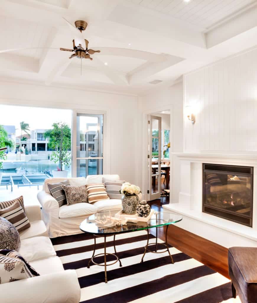 通风的客厅里有条纹地毯和白色沙发上的枕头，旁边有一个玻璃咖啡桌和固定在白色床头板墙上的壁炉。