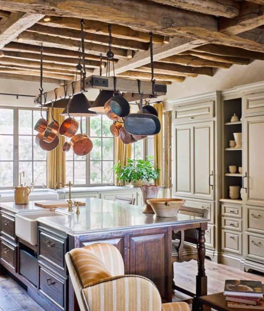 舒适的厨房设有一个配有抽屉的早餐岛和一个带有黄铜固定装置的白色容器水槽，以及一个挂在质朴木梁天花板上的木锅架。