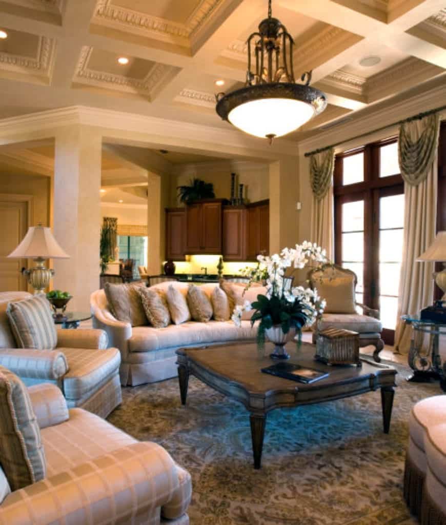 一个巨大的吊灯悬挂在华丽的格子天花板上，照亮了这个客厅，拥有一个木制的咖啡桌，周围是米色的扶手椅和带裙边的沙发，上面摆满了蓬松的枕头。