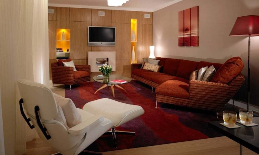 温暖的客厅提供电视之间的嵌入壁龛充满了白色花瓶。它包括一套红色沙发和一张放在小地毯上的白色躺椅。