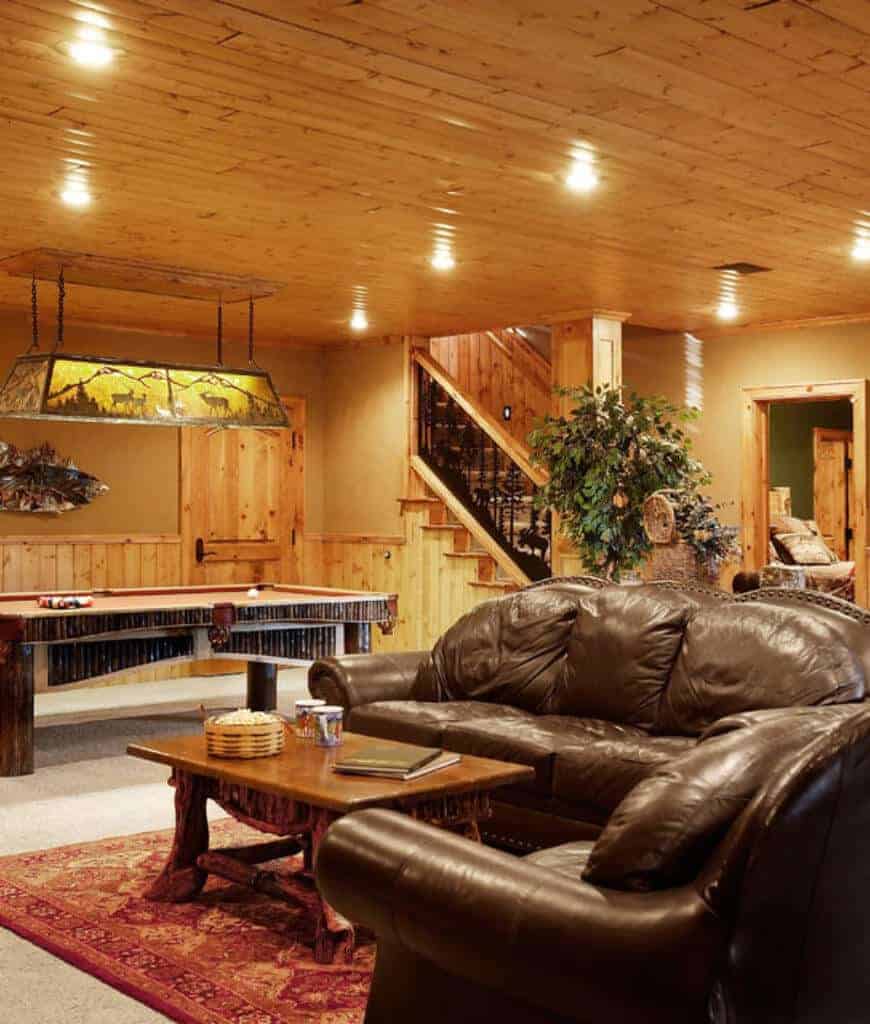 舒适的地下室提供棕色皮革沙发，木质咖啡桌和台球桌在一侧由线性吊灯照明。