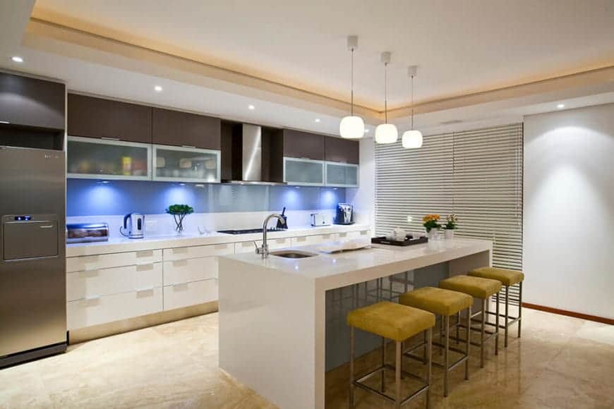 厨房岛台的白色大理石瀑布台面与四个芥末黄色的软垫凳子搭配。这与厨房半岛搭配，具有相同的台面，但通过增强蓝色后挡板的灯光来增强。