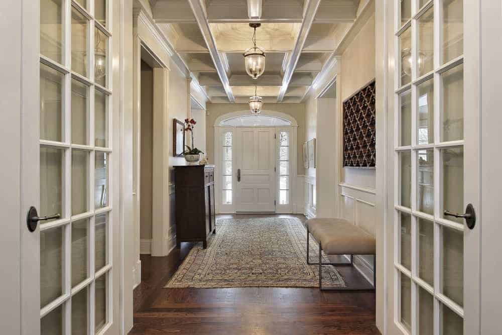 白色的法式法式门通往这间门厅，门厅里铺着硬木地板，上面铺着复古地毯，格子天花板上挂着玻璃吊灯。