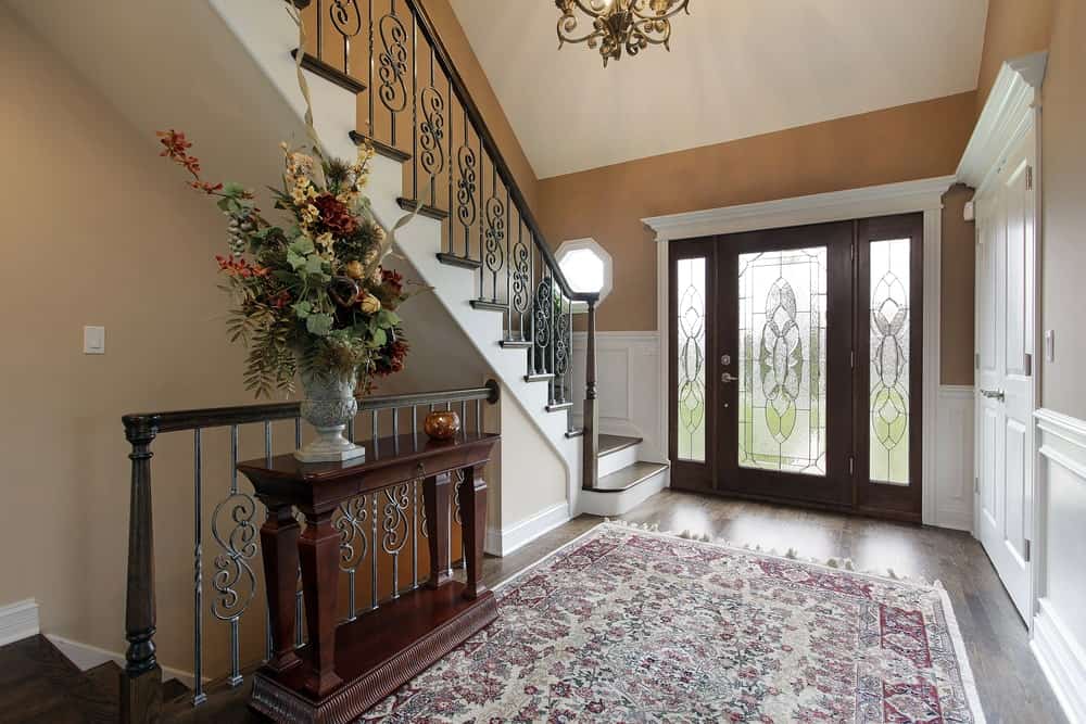 这个门厅的木质正门周围环绕着白色的装饰，在棕色的墙壁和硬木地板的映衬下显得格外突出。地板上铺有花卉图案的区域地毯，从固定在大门上的磨砂玻璃和侧灯中射入的自然光使地毯更加明亮。