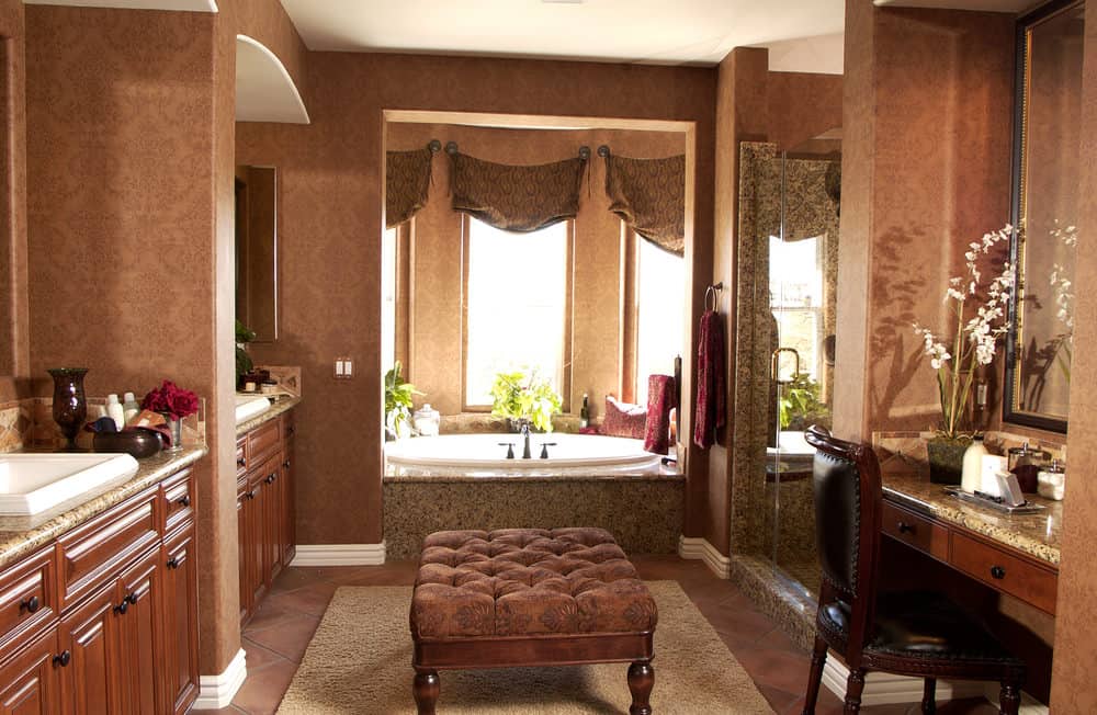 一个棕色的主浴室，有优雅的墙壁和一个优雅的搁脚凳放在中心。屋内有两个盥洗室、一个化妆间和一间步入式淋浴间。