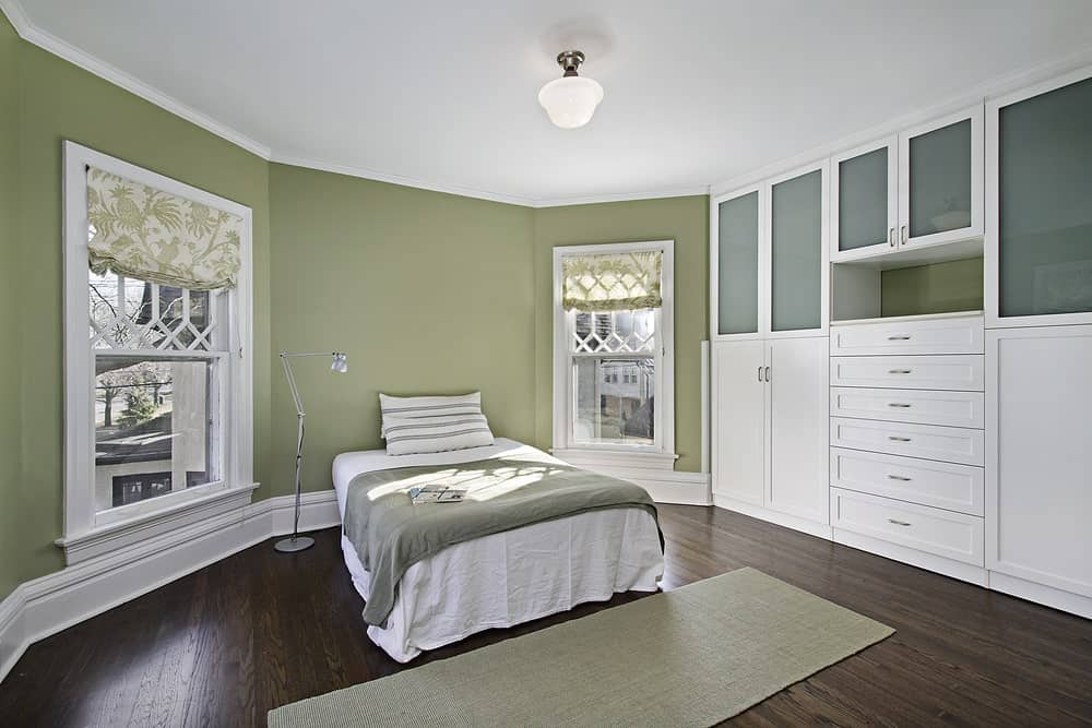 白色的别墅风格的床两侧有两扇高高的窗户，上面有华丽的白色木制设计。深色的木地板与绿色的墙壁形成了很好的互补，与占据一面墙壁的白色嵌入式橱柜和抽屉形成了对比。