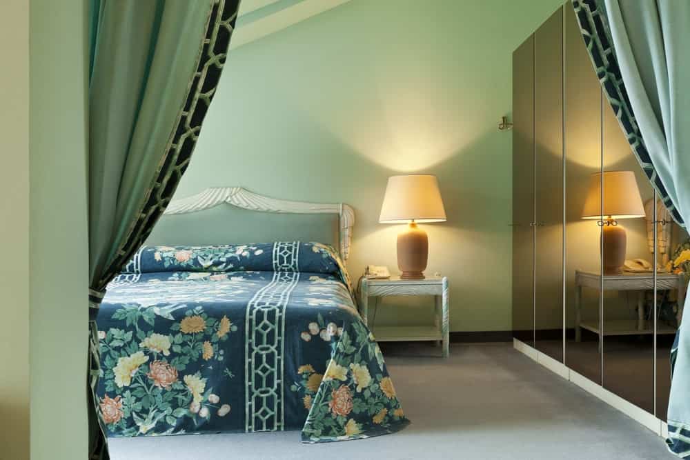 传统的床有一个漂亮的绿色别致的床单，与法式床头板相辅相成，白色床头柜上有一盏台灯。这盏灯在绿色的墙壁上发出温暖的黄色光，与床边光滑的黑色橱柜门形成对比。