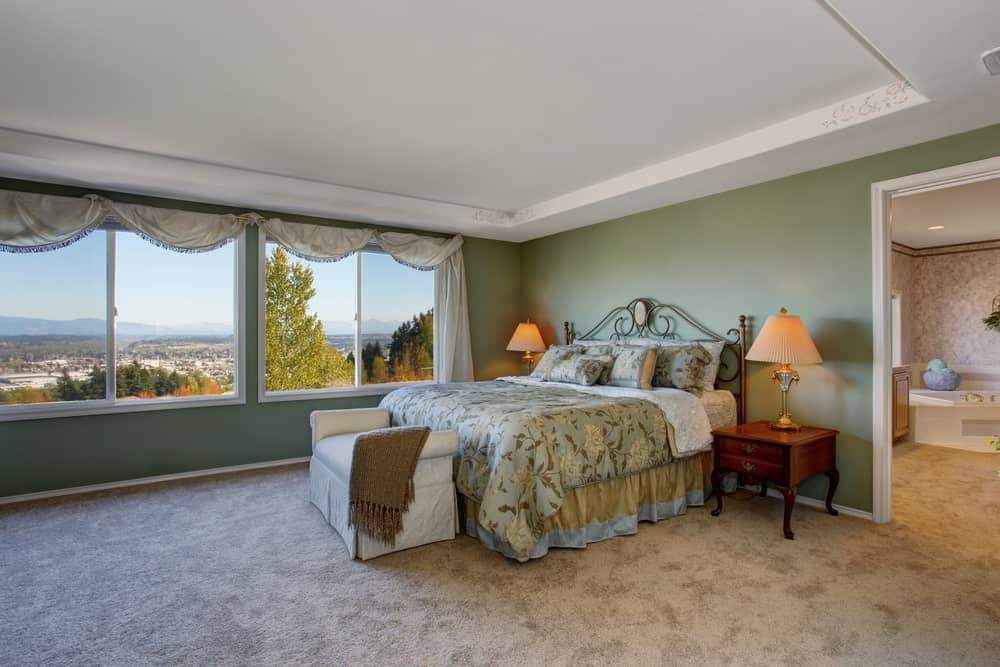 这间绿色墙壁的主卧室通过一对宽大的窗户，与白色托盘天花板和浅灰色地毯地板相匹配，可以欣赏到周围自然的美景。传统的床有一个华丽的铁床头板，与床单的图案相匹配，以达到别致的审美。