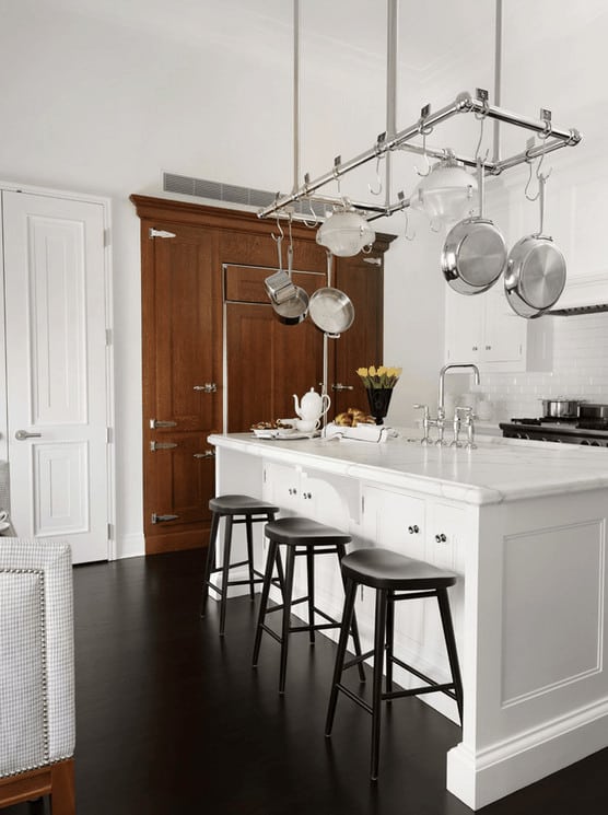 一个木制储物柜在这个白色厨房里很显眼，它有一个不锈钢锅架，挂在早餐岛上，上面有黑色椅子和一个装有铬固定装置的水槽。