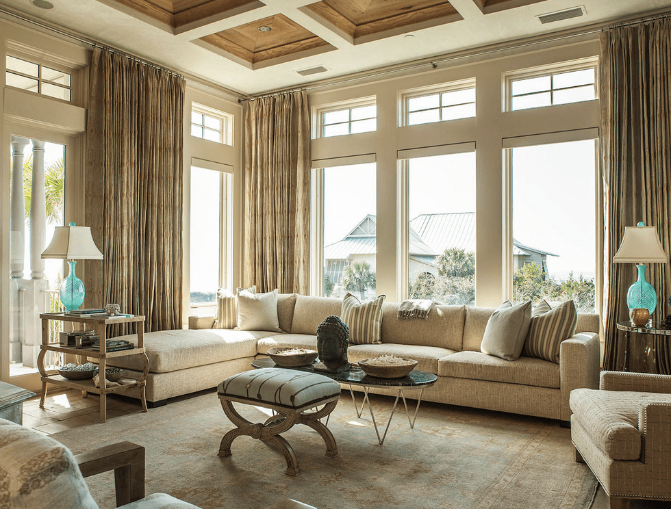 精致的客厅设有木质格子天花板和覆盖米黄色窗帘的玻璃镶板窗户。它有一个金属咖啡桌和l型组合沙发，上面摆满了灰色和条纹枕头。