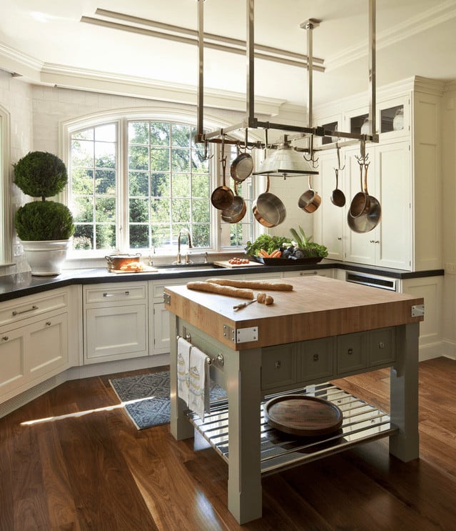 自然光线透过厨房的拱形窗户，厨房里有白色橱柜和金属锅架，悬挂在宽阔的木板地板上的厨房岛台上。