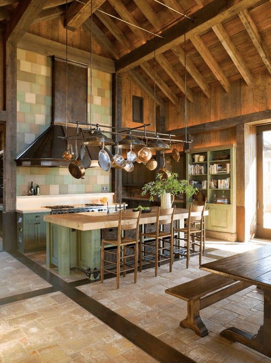 乡村风格的厨房里有绿色的橱柜和岛式吧台，吧台位于教堂高高的天花板上悬挂的金属锅架下面。它包括一个固定在马赛克后挡板瓷砖上的大通风罩。