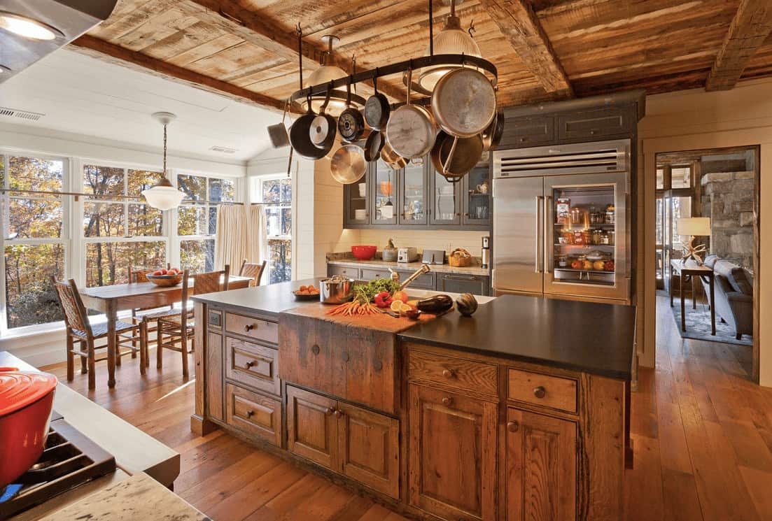 全木制厨房展示灰色橱柜和玻璃前上橱柜，以及金属锅架，挂在天然木质早餐岛上，顶部是黑色花岗岩柜台。