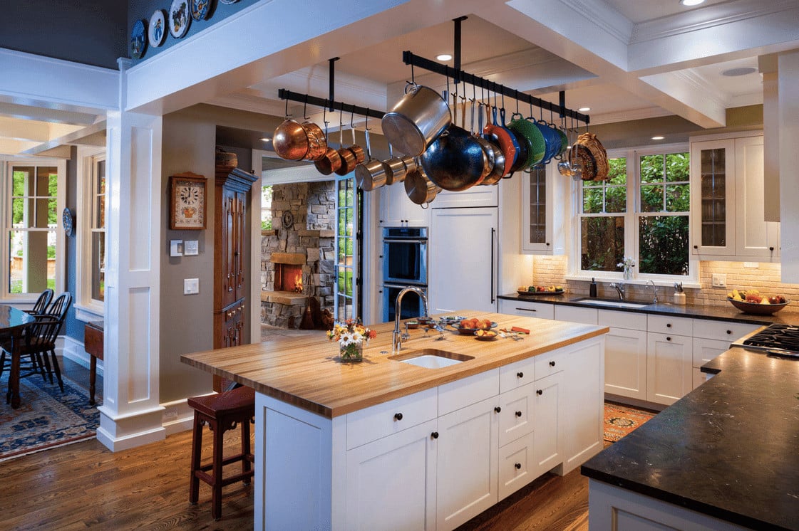 开放式厨房里有白色橱柜，一对线性锅架挂在格子天花板上，上方是木制厨房岛台。